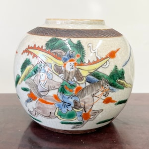 May include: A white ceramic jar with a brown rim and a hand-painted scene of three figures on horseback in a landscape. The figures are painted in shades of blue, green, yellow, and red.