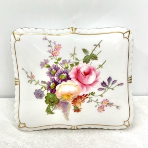 以下が含まれることがあります： 花柄の白い正方形の磁器皿。 皿はスカラップエッジと金色のトリムが特徴です。 花の配置には、ピンクのバラ、紫の花、緑の葉が含まれています。 装飾用の作品です。