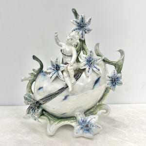 May include: A white ceramic sculpture of a cherub seated on an egg-shaped form, decorated with blue and white flowers and green foliage. The cherub is reaching upwards. The sculpture is a decorative piece.