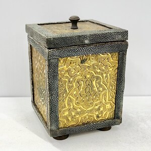 May include: A square decorative box with a gold-coloured, ornate floral design on the front and sides. The box has a dark grey, textured border and a small, round knob on the lid. It rests on four small, round feet.