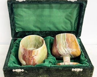 May include: Two polished stone cups with banded patterns in shades of green, brown, and cream, presented inside a rectangular dark green velvet-lined storage box with a hinged lid and silver-toned metal latches on the front edge.
