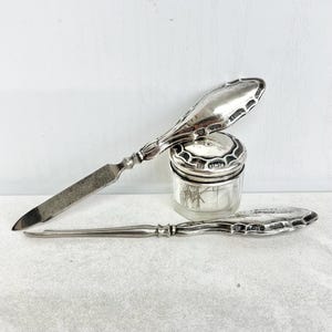 以下が含まれることがあります： 銀製のネイルファイルと銀製のキューティクルプッシャー。どちらも装飾的なハンドルに刻印があり、銀製の蓋が付いた小さな透明なガラス瓶の横に置かれています。瓶の中には数本の細い金属製の道具が入っています。