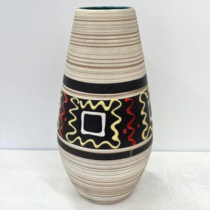 Könnte beinhalten: Eine hohe, dekorative Vase mit cremefarbenem, gestreiftem Körper. Die Vase hat ein schwarzes Band mit einem geometrischen Design in Gelb, Rot und Weiß. Der Rand ist dunkelgrün.