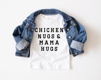 May include: White t-shirt with black text that reads "Chicken Nugs & Mama Hugs" on a white background. The shirt is folded and laid on a white surface with a blue denim jacket draped over it.