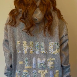 May include: A gray hoodie with the words "CHILD - LIKE FAITH" appliqued on the back in floral patterned fabric. The hoodie has a drawstring hood and long sleeves. The person wearing the hoodie has long, wavy red hair.