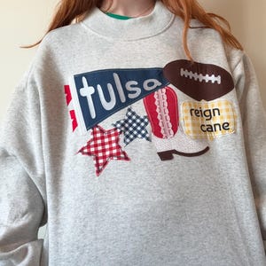 May include: A light gray crewneck sweatshirt featuring a large applique design on the front. The design includes a blue pennant with the text "tulsa", a brown football, a red and white cowboy boot, two gingham stars, and a yellow patch with "reign cane".