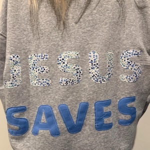 May include: A gray sweatshirt with the words "JESUS SAVES" appliquéd on the back. The letters in "JESUS" are made of a white fabric with a blue floral pattern, while "SAVES" is in solid blue.