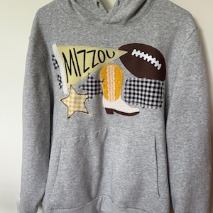 May include: A gray hooded sweatshirt with a front pocket. The sweatshirt features appliqued patches including a football, a cowboy boot, a star, and a pennant with the word "MIZZOU" in yellow and black.
