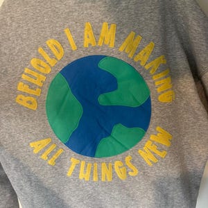 May include: A gray hoodie with a hood and long sleeves. The front features a circular design of the Earth in blue and green, with the words "BEHOLD I AM MAKING ALL THINGS NEW" in yellow lettering around it.