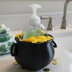 May include: A black pot of gold soap dispenser with a clear soap bottle inside, surrounded by yellow coins. The dispenser sits on a white surface, with green and white shamrock towels in the background. A silver faucet is also visible.