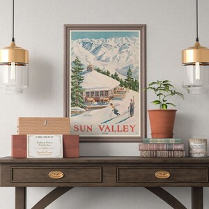 Puede incluir: Un p&oacute;ster vintage con una escena de monta&ntilde;a y una caba&ntilde;a de esqu&iacute;. El p&oacute;ster est&aacute; enmarcado en un marco marr&oacute;n claro y tiene el texto "SUN VALLEY" en letras rojas. El p&oacute;ster est&aacute; colgado en una pared blanca sobre una mesa consola de madera con dos cajones. Hay dos l&aacute;mparas colgantes doradas colgando sobre la mesa.