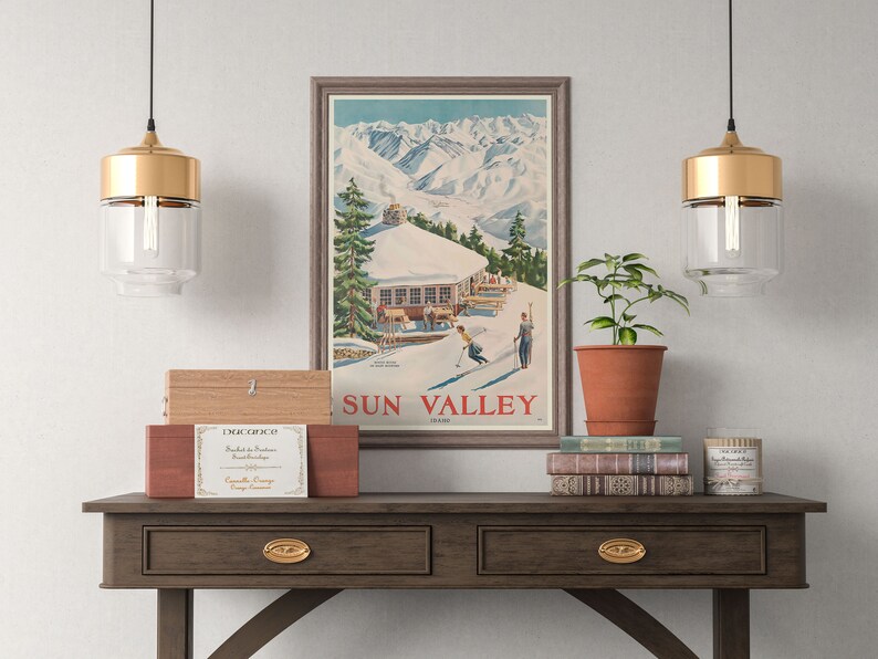 Puede incluir: Un p&oacute;ster vintage con una escena de monta&ntilde;a y una caba&ntilde;a de esqu&iacute;. El p&oacute;ster est&aacute; enmarcado en un marco marr&oacute;n claro y tiene el texto "SUN VALLEY" en letras rojas. El p&oacute;ster est&aacute; colgado en una pared blanca sobre una mesa consola de madera con dos cajones. Hay dos l&aacute;mparas colgantes doradas colgando sobre la mesa.