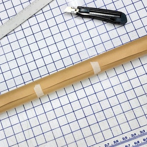 May include: A brown paper roll is taped to itself and laid on a blue grid cutting mat. A utility knife is also on the mat.