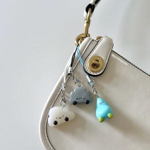 May include: Three whimsical cloud-shaped charms in white, gray, and blue, each with a smiling face, dangle from a white purse. The charms are attached with silver-colored clasps and black cords. The purse has a gold-colored clasp.