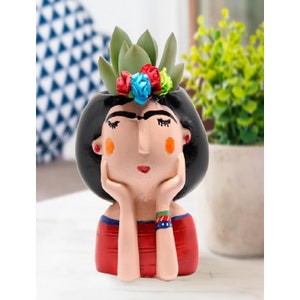 Könnte beinhalten: Ein Keramik-Pflanztopf in Form einer Frau mit schwarzen Haaren, roten Lippen und einer bunten Blumenkrone. Der Topf hat ein rot-weiß gestreiftes Hemd und hält sich mit beiden Händen das Kinn.