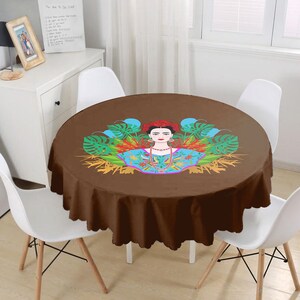 Pode incluir: Toalha de mesa redonda marrom com uma ilustração colorida de Frida Kahlo cercada por folhas e flores tropicais.