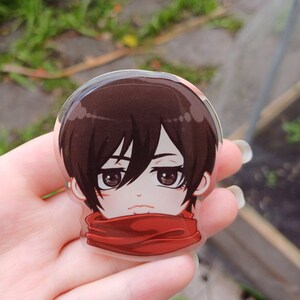 Peut inclure: Un gros plan d'un charme en acrylique transparent repr&eacute;sentant un personnage d'anime aux cheveux bruns, aux yeux rouges et &agrave; une &eacute;charpe rouge. Le personnage porte une &eacute;charpe rouge et a une expression s&eacute;rieuse.
