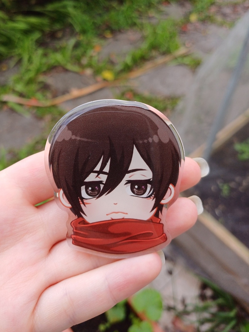 Peut inclure: Un gros plan d'un charme en acrylique transparent repr&eacute;sentant un personnage d'anime aux cheveux bruns, aux yeux rouges et &agrave; une &eacute;charpe rouge. Le personnage porte une &eacute;charpe rouge et a une expression s&eacute;rieuse.