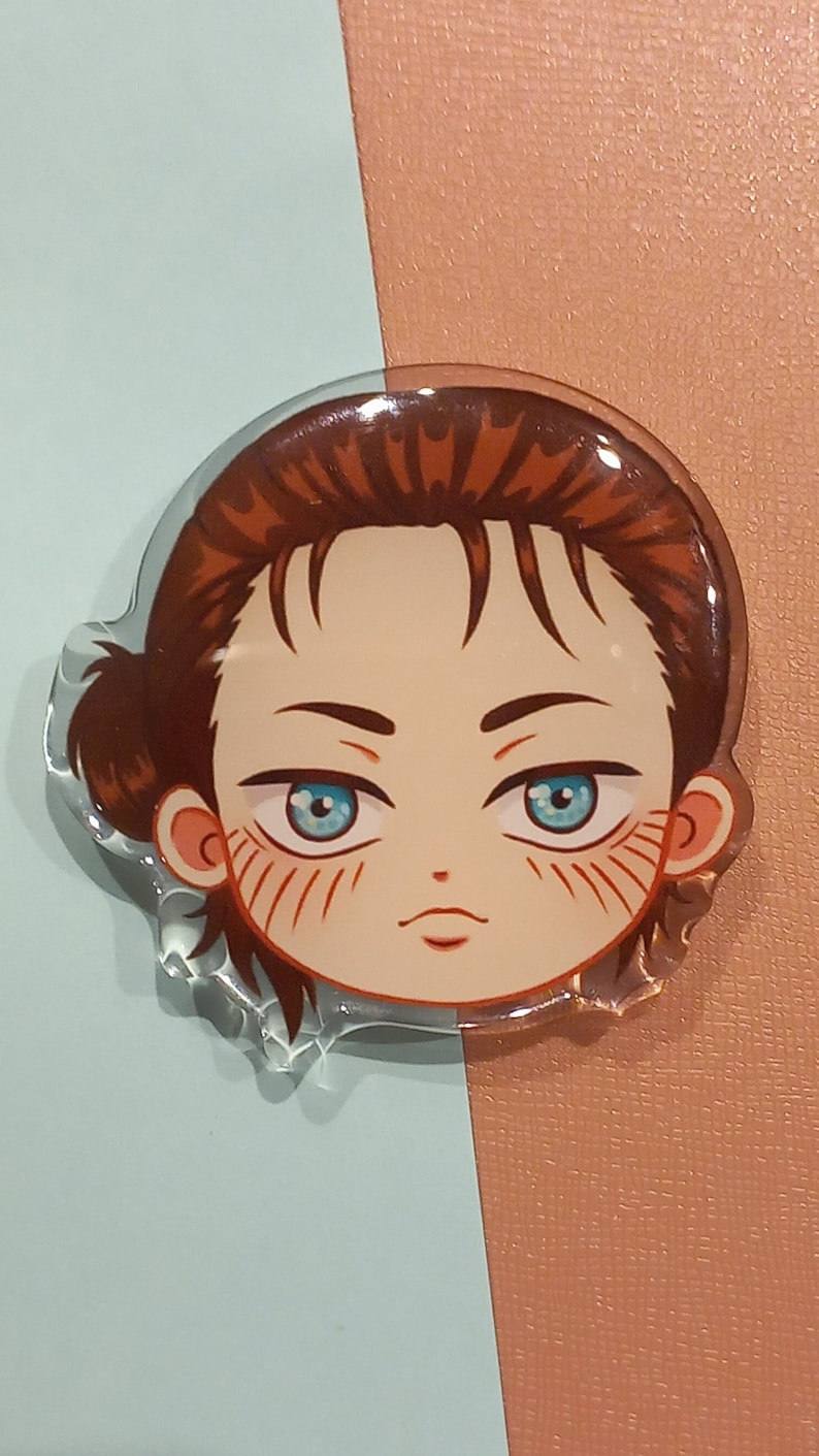 Peut inclure: Un charme en acrylique transparent repr&eacute;sentant un personnage de dessin anim&eacute; avec des cheveux bruns, des yeux bleus et une expression s&eacute;rieuse. Le personnage a un l&eacute;ger blush rose sur les joues.