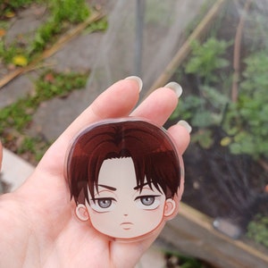 Peut inclure: Un charme rond en acrylique transparent avec une illustration de dessin anim&eacute; d'un jeune homme aux cheveux bruns, aux yeux gris et &agrave; l'expression s&eacute;rieuse. Le charme est tenu dans une main.