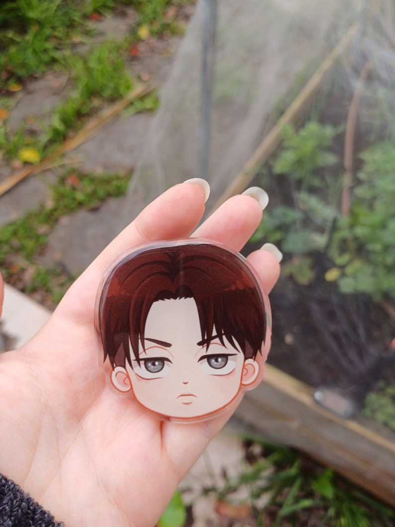 Peut inclure: Un charme rond en acrylique transparent avec une illustration de dessin anim&eacute; d'un jeune homme aux cheveux bruns, aux yeux gris et &agrave; l'expression s&eacute;rieuse. Le charme est tenu dans une main.