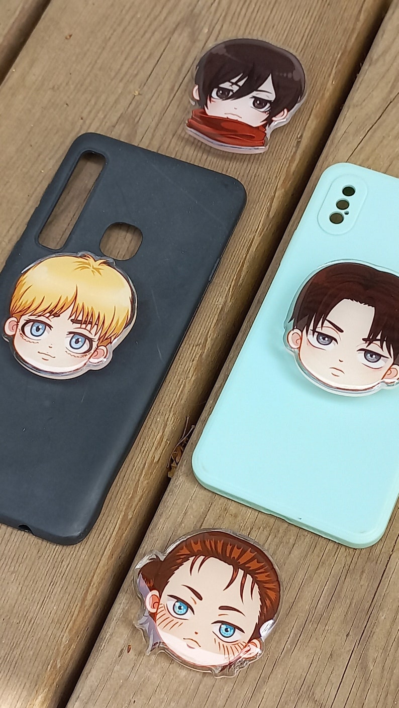 Peut inclure: Quatre poign&eacute;es de t&eacute;l&eacute;phone repr&eacute;sentant des personnages d'anime. Les poign&eacute;es sont en plastique transparent avec des visages de dessins anim&eacute;s color&eacute;s. Les personnages ont les cheveux bruns, blonds et noirs. Les poign&eacute;es sont sur une surface en bois.