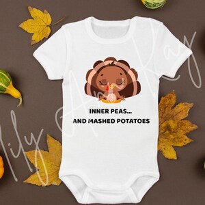 Pode incluir: Body de bebé branco com um desenho de peru de desenho animado. O peru é castanho com um laço rosa e está a meditar. O texto no body diz "INNER PEAS... AND MASHED POTATOES". Folhas de outono e abóboras rodeiam o body.