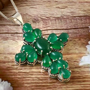 May include: A silver pendant necklace featuring a cross design. The cross is adorned with multiple oval-shaped, vibrant green gemstones. The necklace is displayed on a wooden surface, with white flowers in the background.