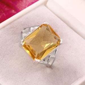 Peut inclure: Une bague en argent avec une grosse pierre jaune rectangulaire. La bague est présentée dans un écrin blanc. La pierre précieuse a une teinte dorée brillante et le corps de la bague a un design géométrique.