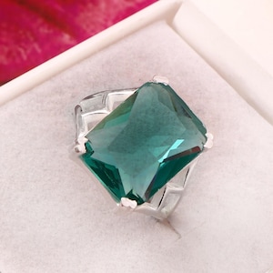May include: A silver ring featuring a large, emerald-cut, teal gemstone. The ring is set in a silver band and presented in a white jewellery box. The gemstone's faceted surface reflects light. The ring is a statement piece.