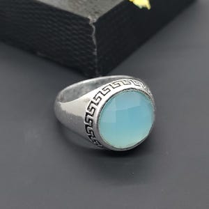 May include: A silver ring featuring a light blue, circular gemstone. The ring is decorated with a Greek key pattern. The ring is displayed on a dark surface.