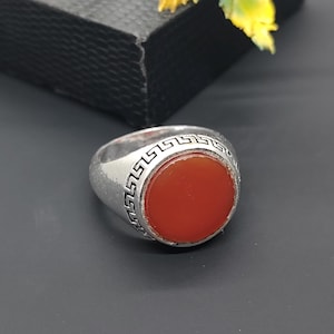 Op de afbeelding: Een zilveren ring met een grote, ronde, oranjerode steen. De ring heeft een getextureerde band met een zich herhalend geometrisch patroon rond de steen. De ring staat tegen een donkere achtergrond.