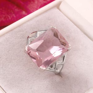May include: A silver ring with a large, square-cut, light pink gemstone. The ring is set in a geometric design and is displayed in a white jewelry box with a soft, white interior. The background features a blurred red floral element.