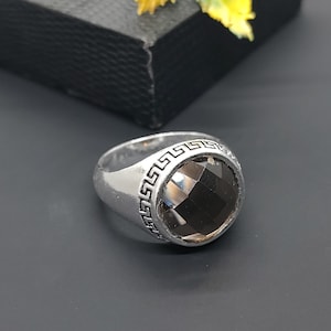 May include: A silver ring with a large, faceted, dark brown gemstone. The ring has a Greek key design around the gemstone. The ring is on a dark gray surface.