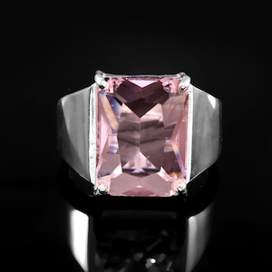 May include: A silver ring featuring a large, rectangular, light pink gemstone. The ring has a wide band and a simple, modern design. The gemstone is faceted, catching the light and creating a sparkling effect. The ring is set against a black background.