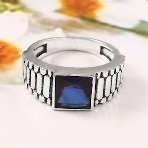 Peut inclure: Bague argentée ornée d'une pierre précieuse carrée bleu foncé. La bague présente un anneau texturé avec un motif de type bracelet de montre. La pierre est sertie dans un cadre carré. L'arrière-plan est flou.