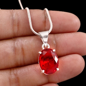 May include: A silver necklace with a vibrant red oval-shaped pendant. The pendant is a faceted gemstone, and the chain is a delicate, textured design. The necklace is displayed against a dark background.