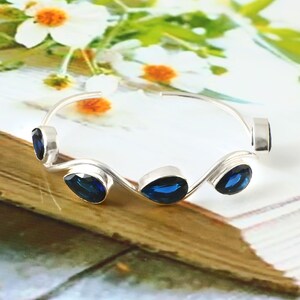 May include: A silver bracelet with five teardrop-shaped, deep blue gemstones. The bracelet has a wavy design and is set against a background of white flowers and a wooden surface. The gemstones are evenly spaced along the bracelet's curve.