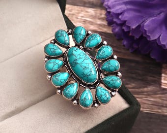 May include: An oval-shaped turquoise ring with a silver-colored metal setting. The ring features a large, central turquoise stone surrounded by smaller, teardrop-shaped turquoise stones. The ring is displayed in a dark green jewelry box.