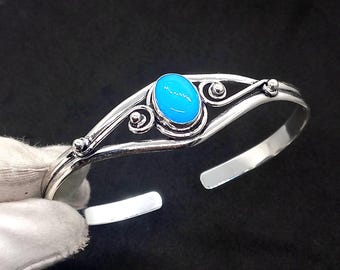 May include: A silver bangle bracelet featuring a blue oval-shaped stone set in the centre. The bracelet has a delicate, ornate design with swirls and small beads.