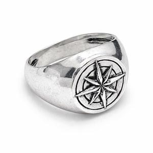 May include: A silver-toned ring with a compass rose design. The ring has a wide band and a raised circular emblem featuring an eight-pointed star. The ring is likely made of metal and has a polished finish.