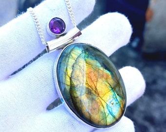 May include: A silver necklace featuring a large, oval labradorite pendant with iridescent blue, green, and yellow hues. A small, round amethyst stone is set above the pendant. The necklace is displayed on a white surface.
