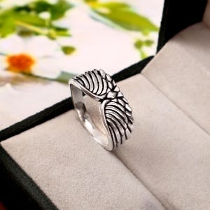 May include: A silver-toned metal ring featuring a detailed design of two symmetrical wings meeting at the centre. The band has an oxidised finish in the recessed areas to highlight the carved feather textures across the surface of the wings.