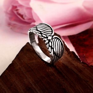May include: A silver-coloured ring featuring a detailed design of two symmetrical wings meeting at the centre. The metal has an oxidised finish to highlight the engraved feather textures. The inner band shows a small 925 stamp indicating the metal purity.