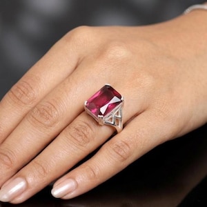May include: A silver ring featuring a large, rectangular, faceted, deep red gemstone. The ring's setting has an open, geometric design on the sides. The gemstone is the focal point, catching the light and showcasing its rich color.