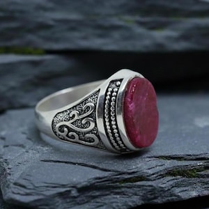 Op de afbeelding: Zilveren ring met een grote, ovaalvormige, dieprode edelsteen. De ring heeft sierlijke details aan de zijkanten en een kralenrand rond de edelsteen. Donkere, getextureerde achtergrond.