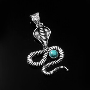 Peut inclure: Pendentif argenté en forme de cobra. Il est orné d'une pierre turquoise et se détache sur un fond noir. La tête du cobra est relevée et son corps est enroulé.
