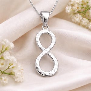 Peut inclure: Un collier avec un pendentif en forme de symbole infini argenté. Le pendentif, à la texture martelée, est suspendu à une fine chaîne. Le collier est présenté sur du satin blanc, avec des fleurs blanches en arrière-plan.