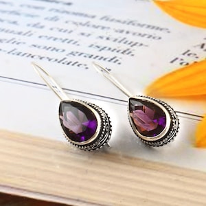May include: A pair of silver teardrop earrings with large, faceted, purple gemstones. The gemstones are set in a decorative, ornate silver setting. The earrings are displayed on a book with text and a yellow flower.