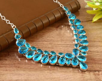 May include: A silver necklace with a series of blue teardrop-shaped gemstones. The gemstones are arranged in a cascading pattern, creating a statement piece. The necklace is displayed on a wooden surface.